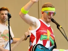 Ando singing into the mic with his guitar on, neon headband, one hand up