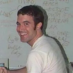 Tom, in a white t-shirt, looking over his shoulder in front of a whiteboard